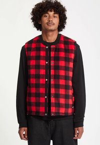 Gilet à carreaux rouge et noir avec boutons-pression et encolure arrondie, fabriqué en tissu doux et texturé, porté par-dessus une chemise noire à manches longues.