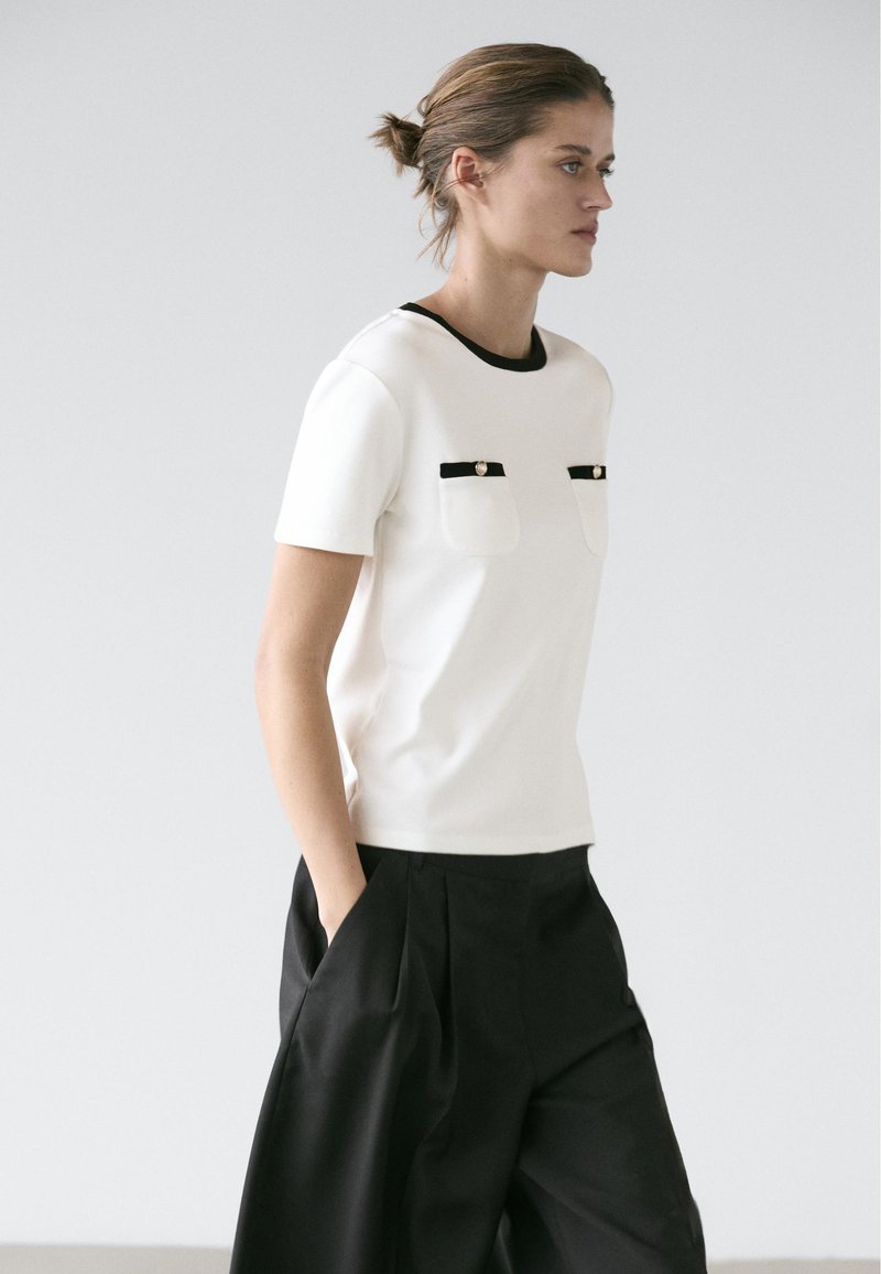 Femme avec les cheveux attachés en chignon, portant un haut blanc à manches courtes avec des poches bordées de noir et un pantalon noir ample, sur un fond clair uni.