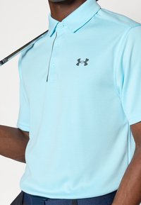 Jasnoniebieska koszulka polo z teksturowanym wzorem, zapięta pod szyją i z ciemnoszarym logo na lewej piersi. Osoba trzyma kij golfowy.