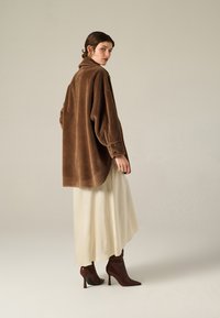 Giacca oversized in pile marrone con colletto alto, dotata di polsini con bottone, abbinata a una gonna di raso color crema e stivaletti marroni.