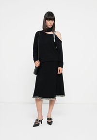 Woman wearing black cold-shoulder sweater with logo strap, black midi skirt with patterned hem, black handbag, and black heeled mules.