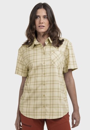 Frau trägt ein beige-kariertes Hemd mit kurzen Ärmeln und rostfarbene Hose, steht vor einem einfarbigen hellen Hintergrund.