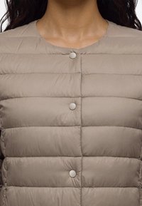 Gros plan d'une personne portant une veste matelassée beige clair avec des boutons-pression ronds et un col arrondi.