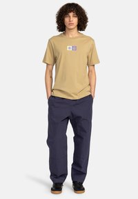 Beige Baumwoll-T-Shirt mit einem kleinen Grafikdesign in der Mitte, kombiniert mit locker sitzenden navyblauen Cargo-Hosen. Schwarze Schuhe mit Gummisohlen.