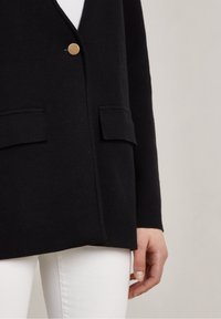 Black knitted cardigan featuring a single gold button, two front pockets, and a relaxed shape. Paired with white pants for contrast.