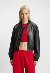 Giacca bomber in pelle nera con chiusura a zip e polsini a costine, indossata sopra un top rosso corto. La modella indossa pantaloni rossi coordinati.