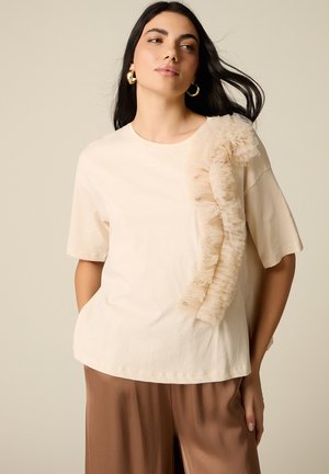 Donna con capelli lunghi e scuri indossa una maglietta beige a maniche corte con dettaglio in tulle arricciato su un lato e pantaloni marroni a gamba larga, in piedi con le mani nelle tasche.