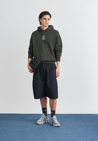 Olivgrüner Hoodie mit Fronttasche und grafischen Details, schwarze knielange Shorts, graue Sportschuhe, stehend auf einem blauen Teppich.