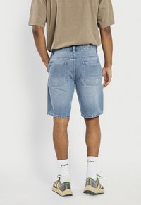 Bărbat purtând pantaloni scurți din denim albastru deschis, tricou bej, șosete albe și adidași bej-gri, stând cu spatele la cameră.