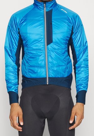 Mężczyzna ubrany w niebieską kurtkę kolarską zapinaną na zamek oraz czarne, wyściełane spodenki kolarskie, stojący z rękami luźno opuszczonymi wzdłuż ciała.