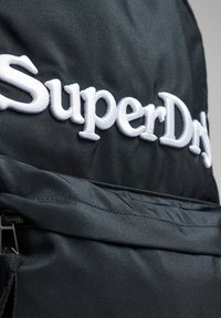 Zaino in tessuto nero con logo "SuperDry" ricamato in bianco, texture liscia e tasca con zip nella parte anteriore.