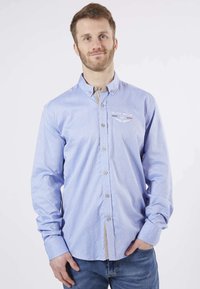 Homme avec une barbe portant une chemise boutonnée à motifs bleu clair et un jean, se tenant avec une main dans la poche devant un fond blanc.