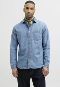 Camicia in denim azzurro chiaro con maniche lunghe, chiusura a bottoni frontale, taschino sul petto e dettagli di cucitura discreti. Abbinata a una sciarpa a fantasia.