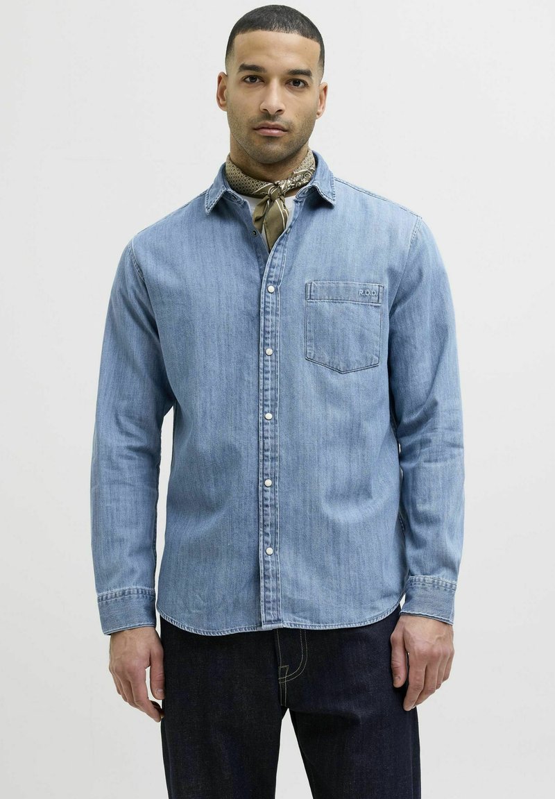Camicia in denim azzurro chiaro con maniche lunghe, chiusura a bottoni frontale, taschino sul petto e dettagli di cucitura discreti. Abbinata a una sciarpa a fantasia.