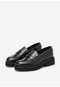 Par de mocasines de cuero negro con suelas gruesas y tracción, y costura tipo moc en un fondo gris claro.
