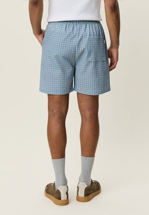 Mann trägt hellblaue karierte Shorts, graue Socken und braune Sneakers, steht mit den Händen in den Taschen, von hinten gezeigt.