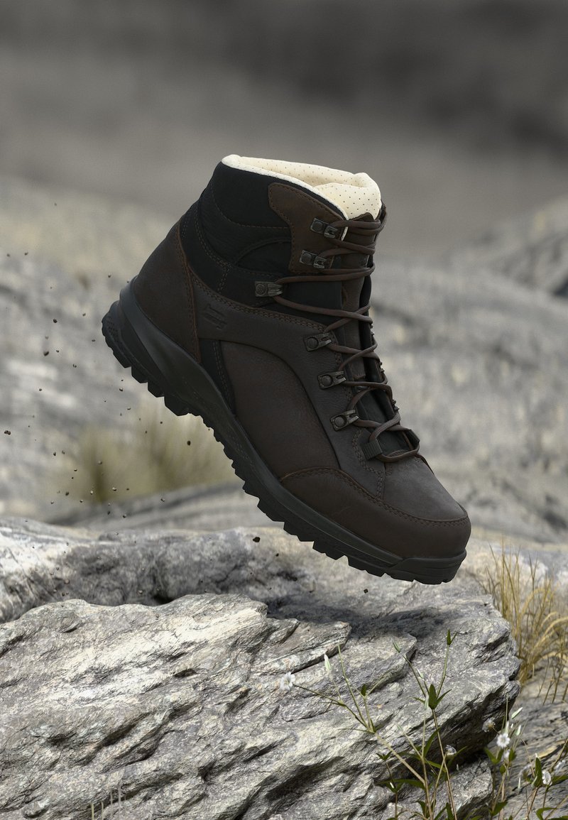 Σκούρο καφέ πεζοπορικό μποτάκι με παχιά σόλα και κορδόνια, κρεμασμένο πάνω από βραχώδες έδαφος με αραιό γρασίδι και θολό φυσικό φόντο.