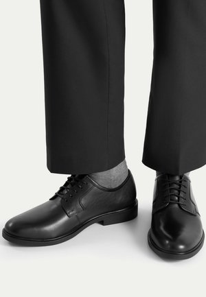 Czarne skórzane wizytowe buty sznurowane noszone z szarymi skarpetkami i czarnymi spodniami na jednolitym białym tle.
