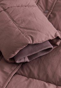 Prošívaná bunda v mauve barvě s hladkým, texturovaným vnějším materiálem a složenou vnitřní pletenou vrstvou. Obsahuje prošívaný vzor a knoflíky.
