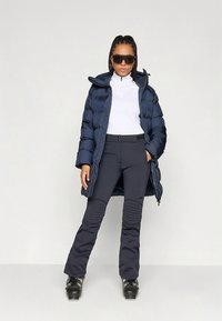 Granatowa kurtka puchowa z poziomym pikowaniem, zestawiona z dopasowanymi granatowymi spodniami z teksturowanymi wstawkami na kolanach oraz czarnymi butami narciarskimi. Biały top z wysokim kołnierzem.