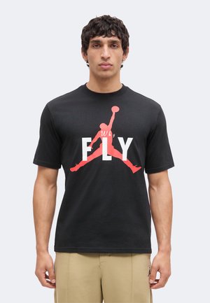 Férfi áll szemben, fekete pólót visel, amelyen piros kosárlabda sziluett és "Why FLY" felirat van, bézs nadrág.