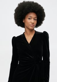 Ženska z naravno afro frizuro v črni žametni obleki s dolgimi rokavi na enobarvni podlagi.
