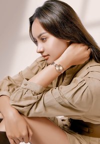 Reloj cuadrado de color marrón dorado con una correa de metal y esfera redonda, mostrado en una muñeca, acompañado de una blusa beige texturizada.