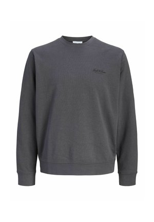 Garçon blond souriant, portant un sweat-shirt gris foncé à manches longues, un pantalon noir ample et des baskets noires, debout sur un fond clair uni.