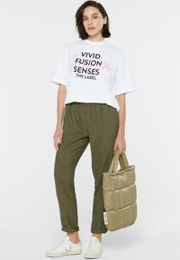 White graphic t-shirt with black and pink text, olive green elastic-waist pants, light-colored sneakers, and a padded olive tote bag.