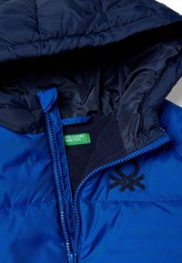 Niebieska kurtka pikowana z przeszyciami, kołnierzem podszytym polarem, logo oraz zieloną metką marki. Zapinana na suwak z dołączonym kapturem.