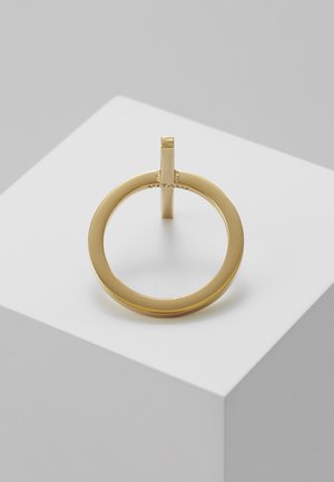 Minimalistischer Goldring mit einem kleinen vertikalen Balken, dargestellt auf einem weißen kubischen Podest vor einem hellgrauen Hintergrund.