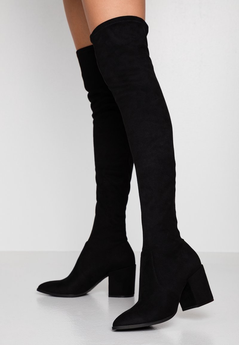 Bottes noires en daim jusqu'au genou avec un bout pointu et un talon large, offrant une texture lisse et un design ajusté, mettant en valeur la jambe.