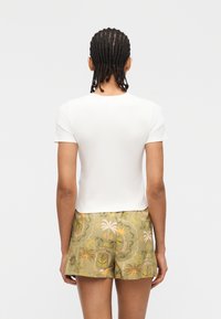 Άτομο με πλεξούδες, που φοράει λευκό κοντομάνικο μπλουζάκι και σορτς σε ελιά πράσινο χρώμα με τροπικό σχέδιο από φοινικόδεντρα, η εικόνα από πίσω.