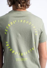 Olijfgroene T-shirt van zachte katoen met een ronde gele grafische tekstontwerp op de achterkant, inclusief merknaam en projectdetails.