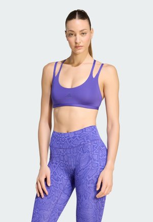 Kvinde står iført lilla sports-bh med dobbelte stropper og lilla mønstrede leggings med høj talje mod en ensfarvet baggrund.