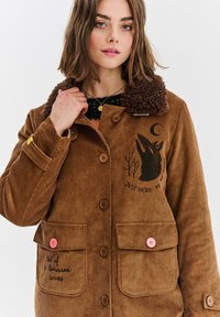 Brązowa kurtka z korduroyu z brązowym puszystym kołnierzem, z dwoma przednimi kieszeniami, guzikami oraz czarnymi haftowanymi grafikami i tekstem.