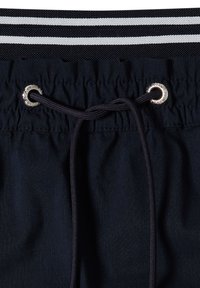 Taille élastique avec cordon noir passant à travers des œillets métalliques argentés sur un tissu foncé sous une bande rayée noire et blanche.