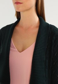 Tmavě zelený pletený kardigán s žebrovaným límcem, nošený přes světle růžový top s výstřihem do V. Viditelná textura a detaily vrstveného designu.