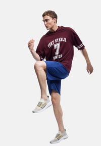Bordeaux hoodie met de tekst "VERY STABLE 7 GENUIS", gecombineerd met blauwe shorts en lichtgrijze sportschoenen met zwarte accenten en een patroon in de veters.