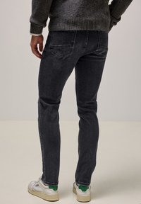 Jeans slim fit de color gris oscuro con dos bolsillos traseros, tela sutilmente texturizada y diseño de tobillo ajustado, llevados con zapatos blancos con acentos verdes.