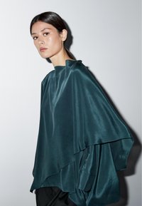 Teal satin poncho med asymmetrisk drapering, som har hög hals och flödande silhuett mot en enfärgad bakgrund.
