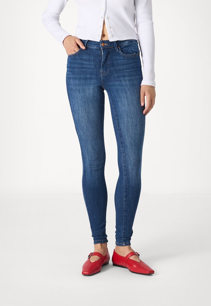 Jeans i mörkblå denim med en slät yta och en klassisk design med fem fickor. Matchas med röda platta skor med utskärningar.