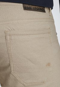 Pantalons beiges texturés présentant un motif tissé, avec une étiquette en cuir "PME LEGEND" et des coutures visibles sur les poches arrière.