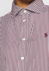 Camisa de botones a rayas en burdeos y blanco. Presenta un cuello clásico y botones blancos, con un pequeño detalle de logo en el pecho.