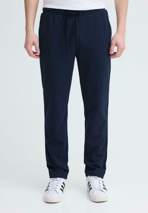 Hombre vistiendo pantalones deportivos azul marino con cordón negro y zapatillas blancas con rayas negras sobre fondo blanco.