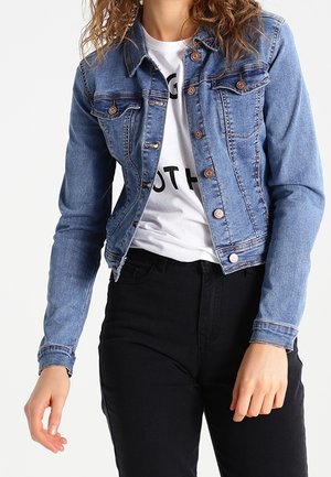Vrouw draagt een blauwe spijkerjas over een wit bedrukt t-shirt en een zwarte broek, staand tegen een witte achtergrond.