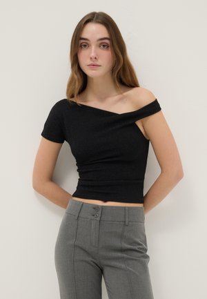 Mujer joven con cabello largo, vistiendo una blusa negra asimétrica de un solo hombro y pantalones grises de talle alto, de pie contra una pared clara.