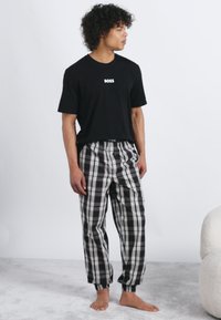 Tânăr în picioarele goale, purtând un tricou negru "BOSS" și pantaloni de pijama în carouri alb-negru, stând pe un covor gri deschis lângă un scaun alb.