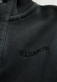 Juodas medvilnės džemperis su ketvirčio užtrauktuku, raukiniais apykakle ir subtiliu siuvinėtumu „ALLSAINTS“ netoli apvado. Minkšta tekstūra.