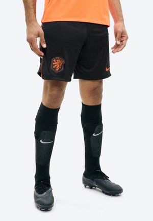 Mužský futbalista v čiernych šortkách s erbom tímu Holandska, čiernymi chráničmi holení Nike, čiernymi kopačkami a oranžovým dresom.